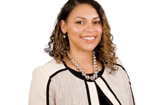 Woman with shoulder-length curly hair, wearing a cream and black jacket with a pearl necklace, smiling in front of a white background.
