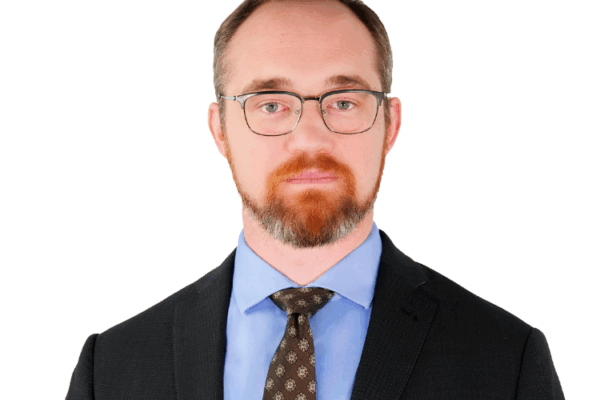 Man with short hair and a beard, wearing glasses, a dark suit, light blue shirt, and patterned tie, facing forward with a neutral expression.
