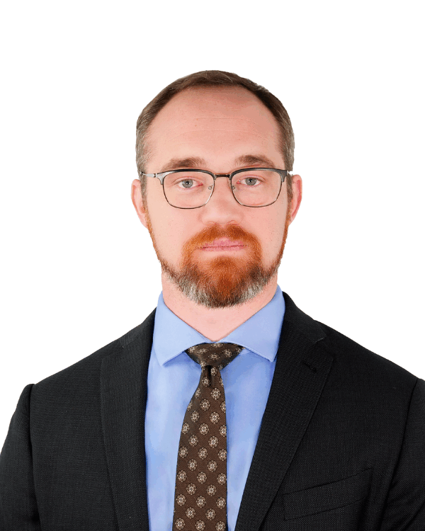 Man with short hair and a beard, wearing glasses, a dark suit, light blue shirt, and patterned tie, facing forward with a neutral expression.