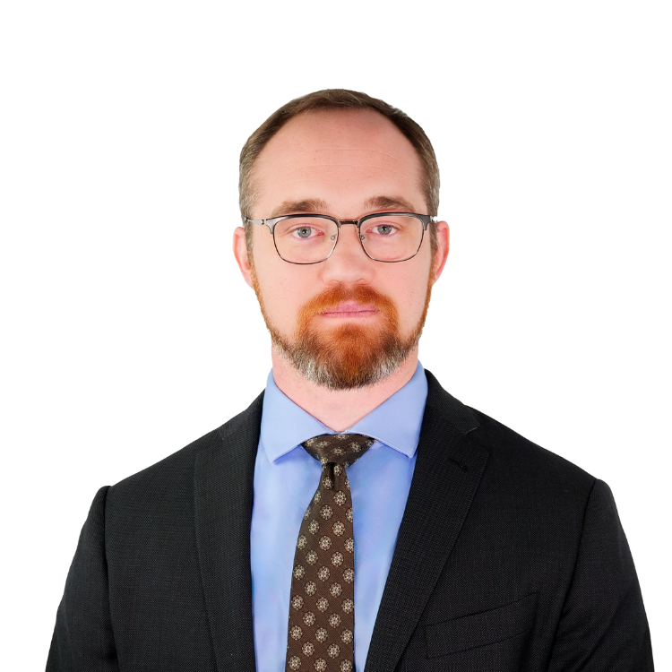 Man with short hair and a beard, wearing glasses, a dark suit, light blue shirt, and patterned tie, facing forward with a neutral expression.