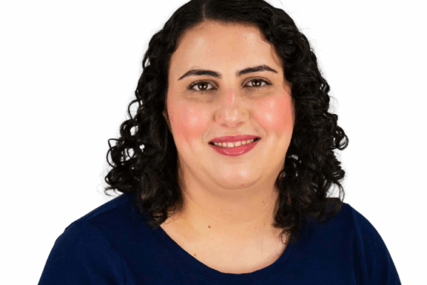 Woman with shoulder-length dark curly hair, wearing a navy blue top, smiling gently in front of a white background.