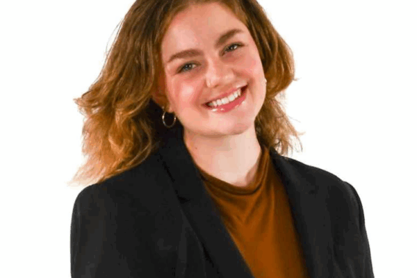Woman with shoulder-length wavy hair, wearing hoop earrings, a rust-colored top, and a black blazer, smiling in front of a white background.