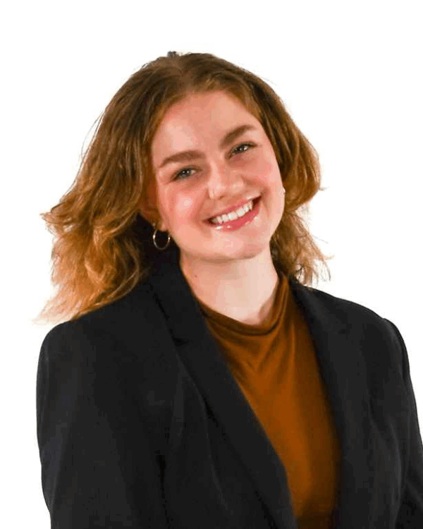 Woman with shoulder-length wavy hair, wearing hoop earrings, a rust-colored top, and a black blazer, smiling in front of a white background.