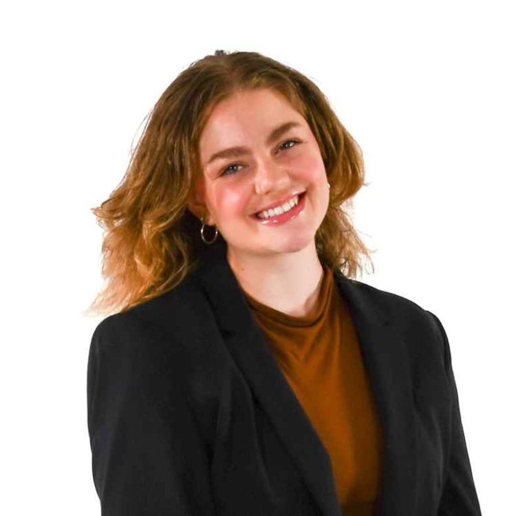 Woman with shoulder-length wavy hair, wearing hoop earrings, a rust-colored top, and a black blazer, smiling in front of a white background.