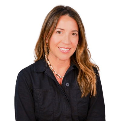 Woman with long wavy hair, wearing a black zip-up jacket and a chunky chain necklace, smiling against a white background.