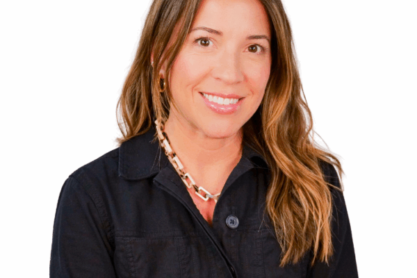 Woman with long wavy hair, wearing a black zip-up jacket and a chunky chain necklace, smiling against a white background.
