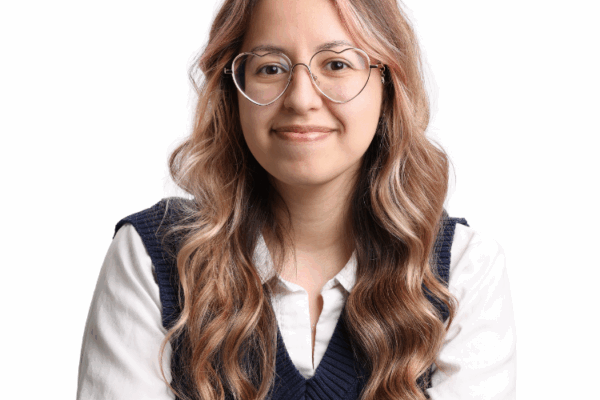 Woman with long wavy hair and light pink highlights, wearing large heart-shaped glasses, a white blouse, and a navy sweater vest, smiling in front of a white background.