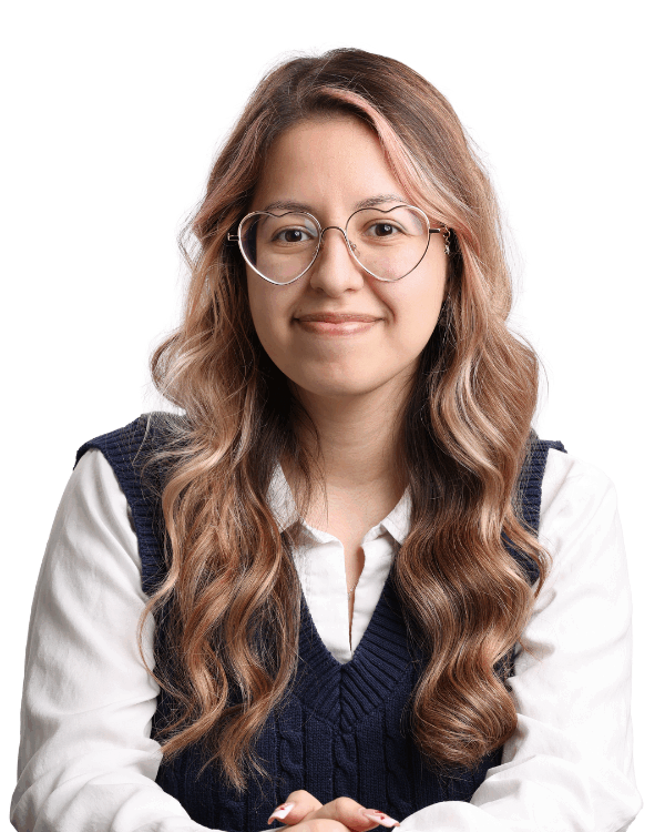 Woman with long wavy hair and light pink highlights, wearing large heart-shaped glasses, a white blouse, and a navy sweater vest, smiling in front of a white background.