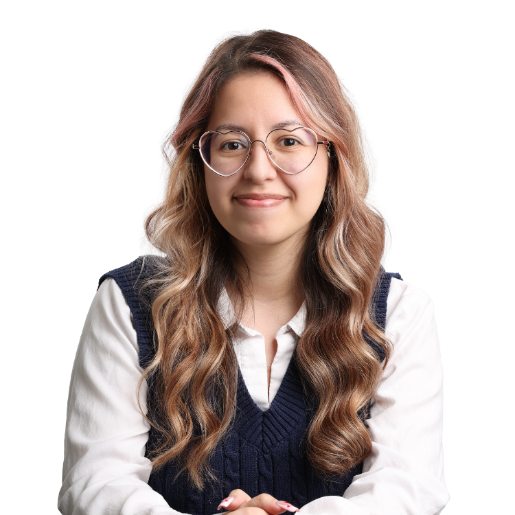 Woman with long wavy hair and light pink highlights, wearing large heart-shaped glasses, a white blouse, and a navy sweater vest, smiling in front of a white background.