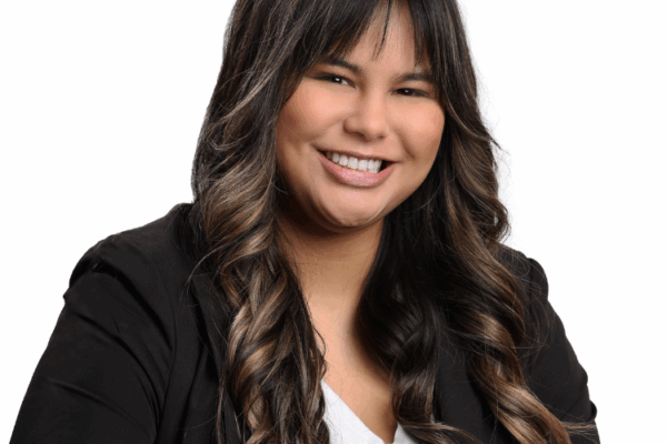 Woman with long, curled dark brown hair and bangs, wearing a black blazer and white blouse, smiling brightly in front of a white background.