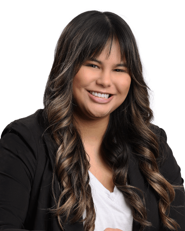 Woman with long, curled dark brown hair and bangs, wearing a black blazer and white blouse, smiling brightly in front of a white background.