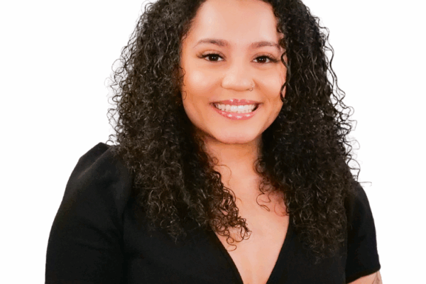 Woman with medium-length curly hair, wearing a black top and showing tattoos on her arm, smiling warmly in front of a white background.