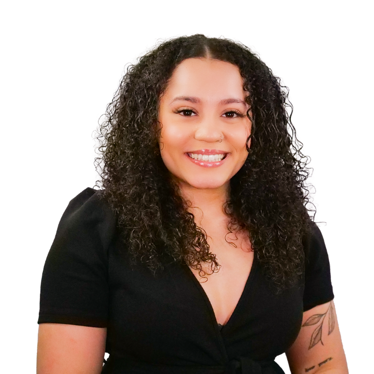Woman with medium-length curly hair, wearing a black top and showing tattoos on her arm, smiling warmly in front of a white background.