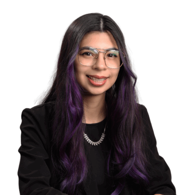 Woman with long dark hair dyed purple at the ends, wearing glasses, a black top, and a silver necklace, smiling gently in front of a white background.