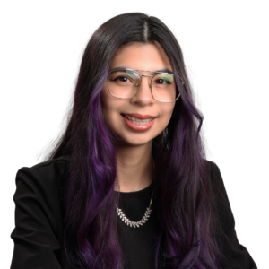 Woman with long dark hair dyed purple at the ends, wearing glasses, a black top, and a silver necklace, smiling gently in front of a white background.