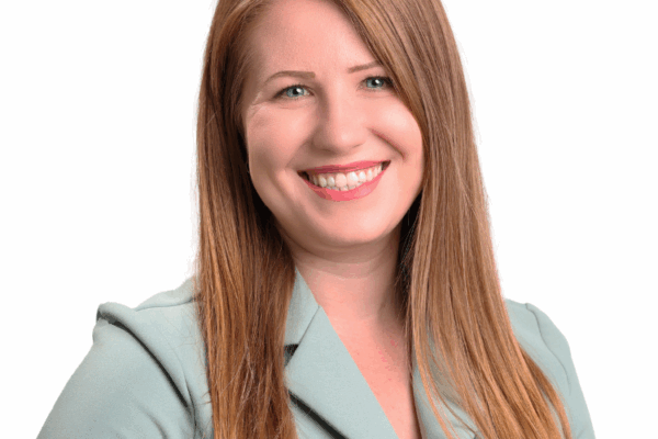 Woman with long straight red-blonde hair, wearing a light green blazer, smiling confidently in front of a white background.