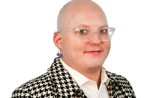 Bald person with clear glasses and a bold earring, wearing a black and white houndstooth jacket over a white shirt, smiling slightly.
