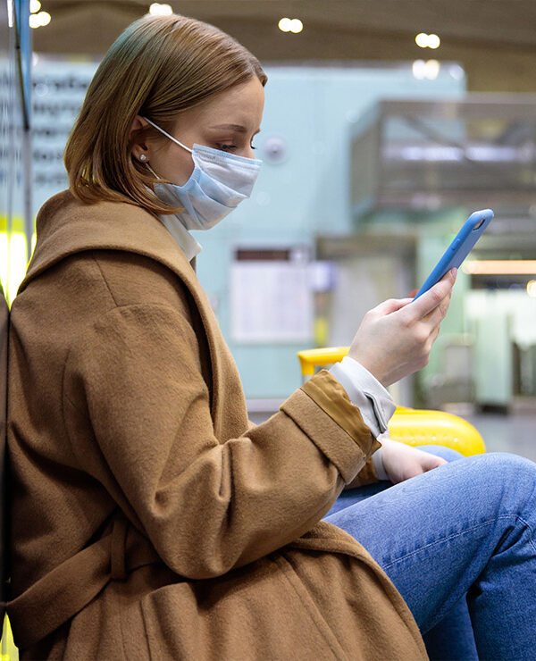 A woman wearing a face mask sits at an airport while scrolling through her phone.