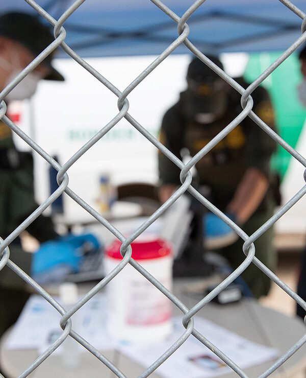 CBP agents wearing PPE processing someone wearing a mask behind a chain-link fence.