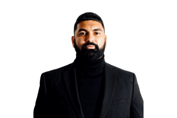 A man with a neatly trimmed beard and short hair, wearing a black turtleneck and a black blazer, stands against a plain white background.