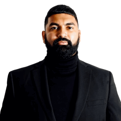 A man with a neatly trimmed beard and short hair, wearing a black turtleneck and a black blazer, stands against a plain white background.