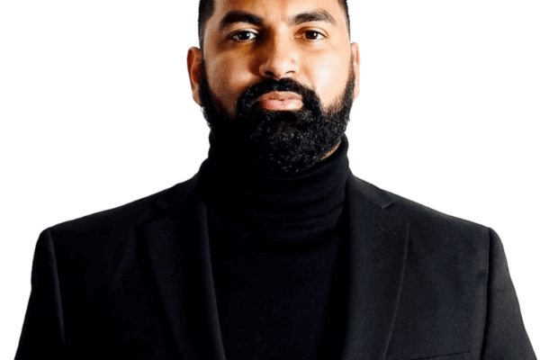 A man with a neatly trimmed beard and short hair, wearing a black turtleneck and a black blazer, stands against a plain white background.