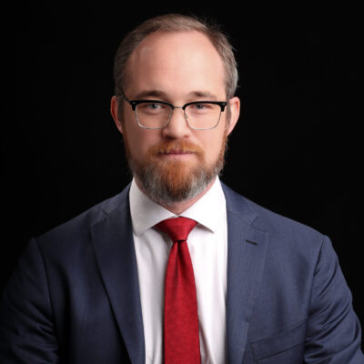 Headshot of Chris. He is a white man with glasses. He is wearing a navy blazer, white collared shirt, and a red tie.