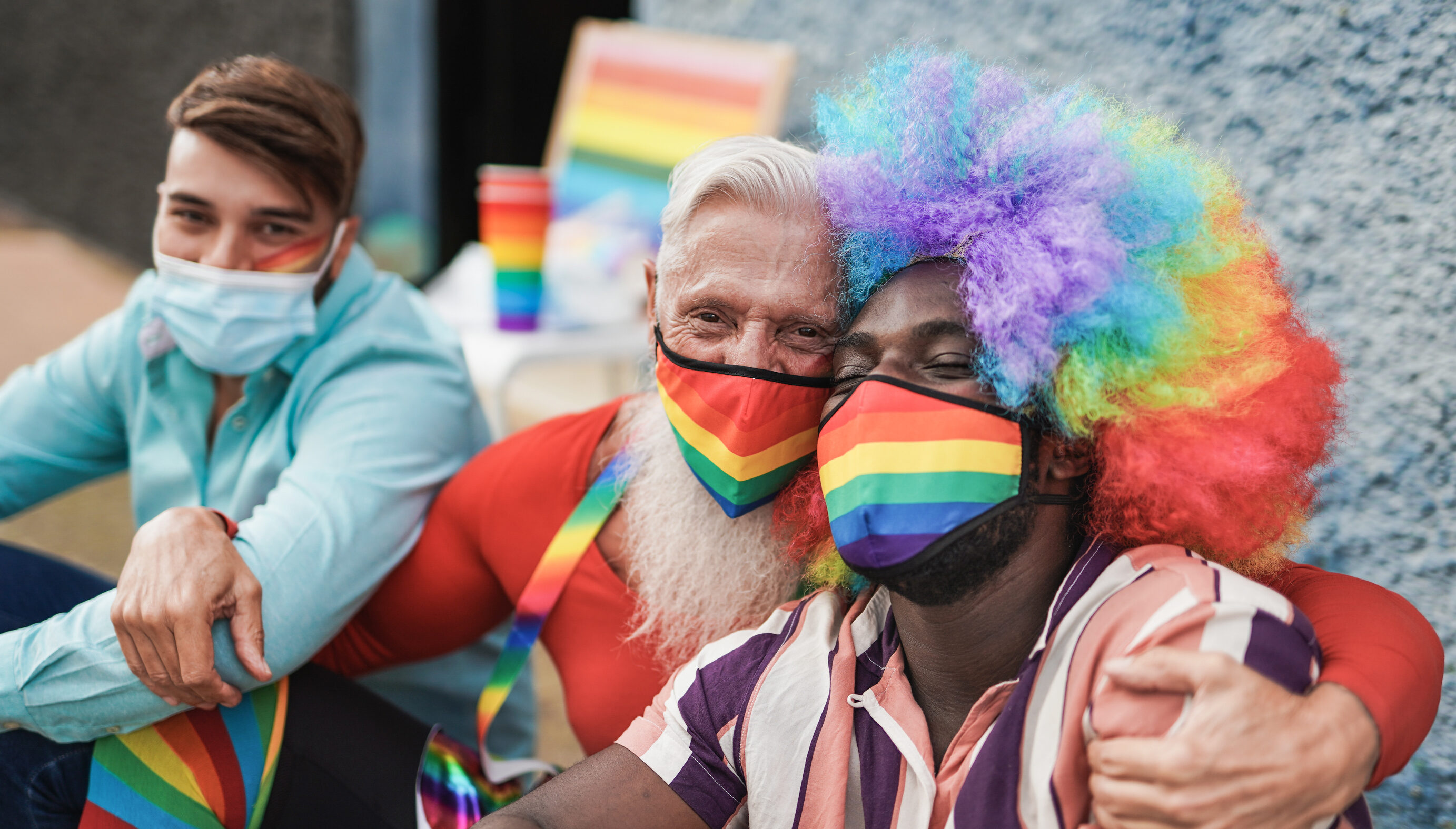 Three men at a LGBTQ+ parade. They are sitting on the curb. The man on the right is a black man with a rainbow afro wig on and a rainbow mask. He is being hugged by the man to his left who also has a rainbow mask on. The third man has a blue mask on.