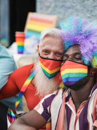 Three men at a LGBTQ+ parade. They are sitting on the curb. The man on the right is a black man with a rainbow afro wig on and a rainbow mask. He is being hugged by the man to his left who also has a rainbow mask on. The third man has a blue mask on.