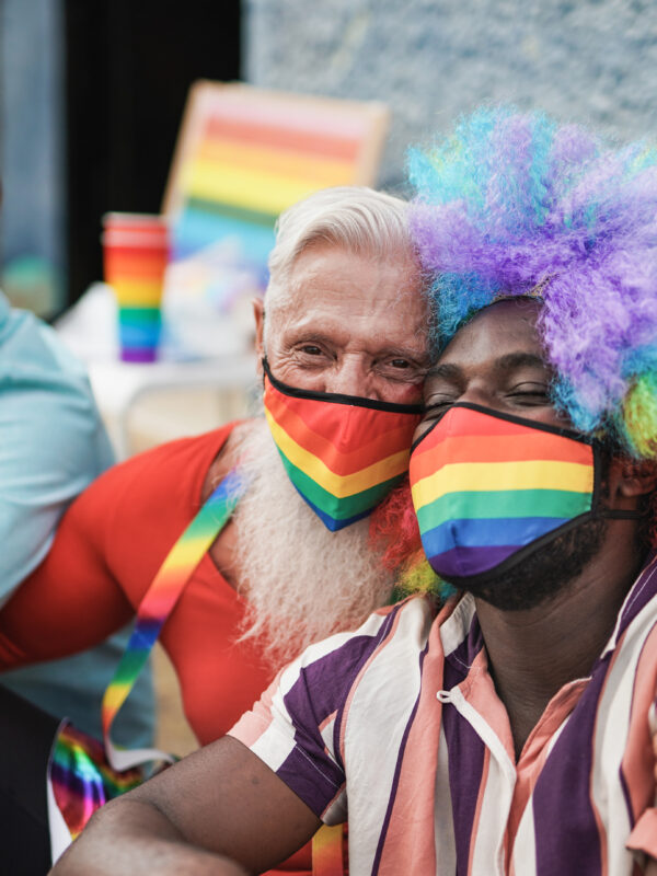 Three men at a LGBTQ+ parade. They are sitting on the curb. The man on the right is a black man with a rainbow afro wig on and a rainbow mask. He is being hugged by the man to his left who also has a rainbow mask on. The third man has a blue mask on.