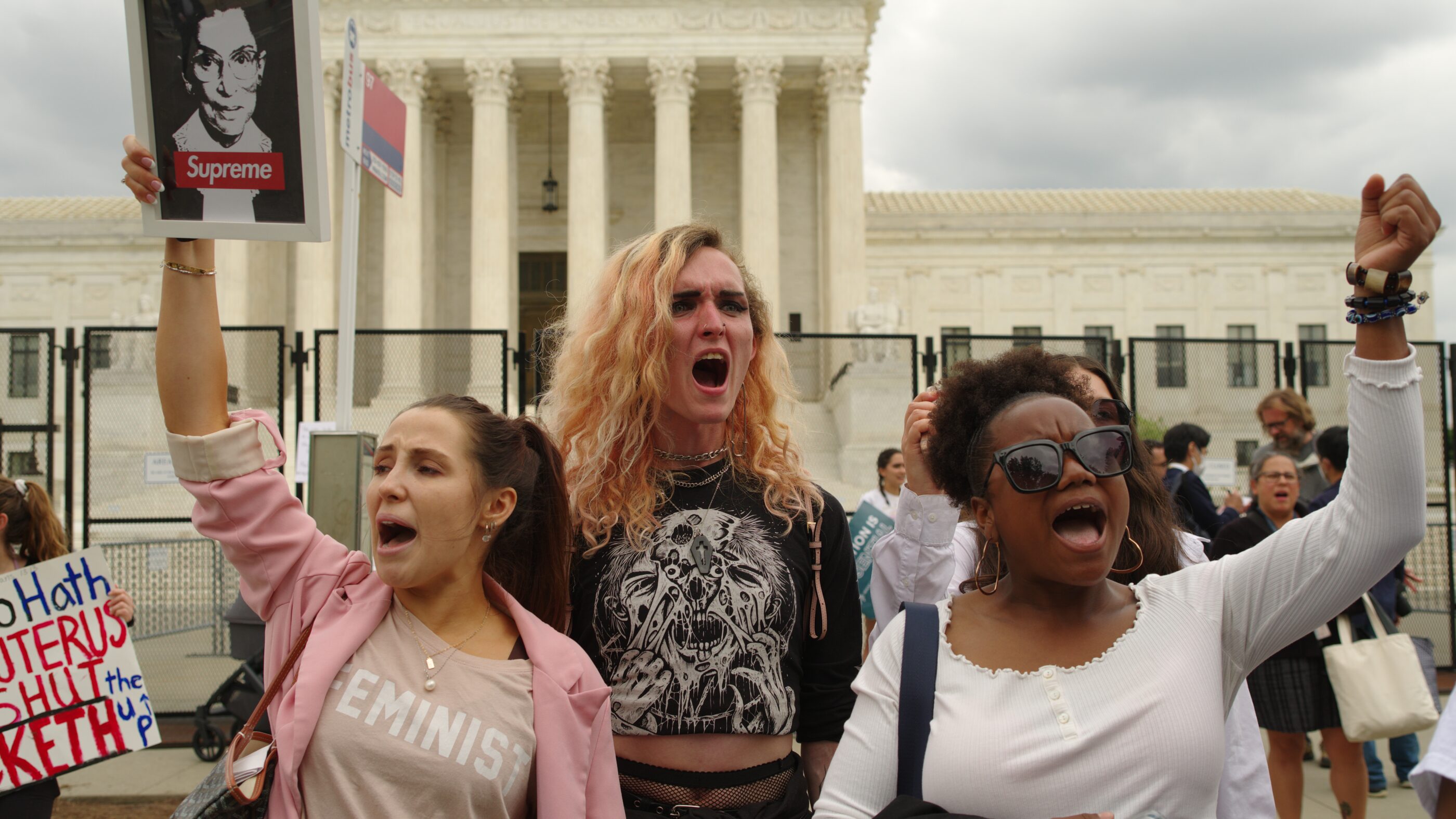 Three people in front of Supreme Court Building. Their fists are in the air and one is holding a picture of RBG.
