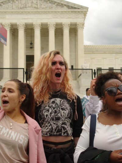 Three people in front of Supreme Court Building. Their fists are in the air and one is holding a picture of RBG.