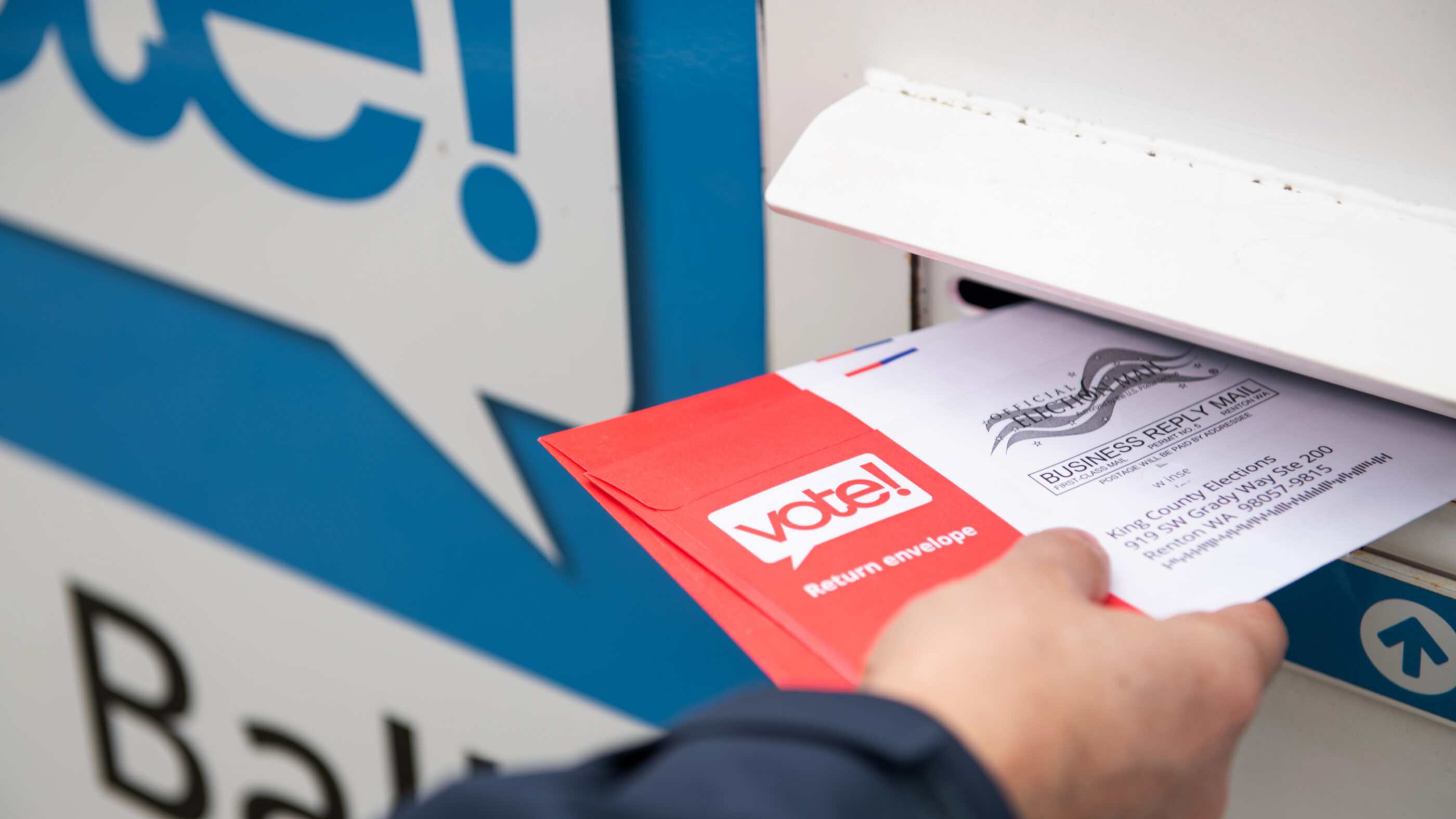 A hand holding a ballot that is half way in the slot of a ballot box.