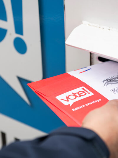 A hand holding a ballot that is half way in the slot of a ballot box.