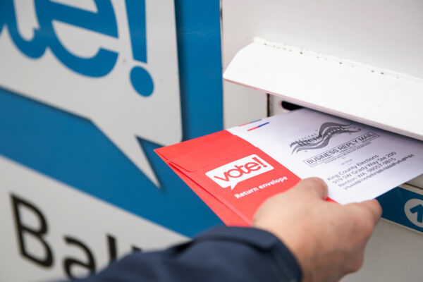 A hand holding a ballot that is half way in the slot of a ballot box.