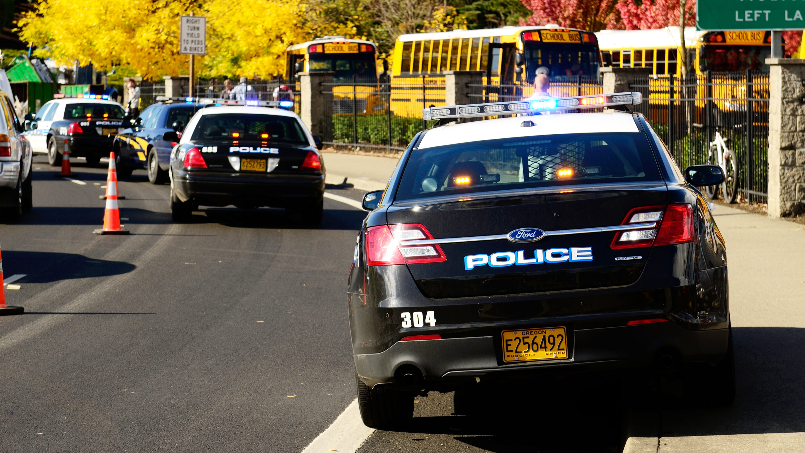 Line of police cars parked in front of school. There is also a line of school buses.
