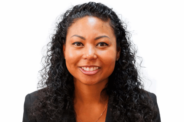A woman with medium length black curly hair wearing a Black blazer and gold necklace smiling against a white background.