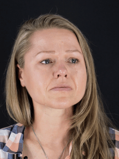 Client, Rebecca Leavitt. She is a white woman with blonde hair, blue eyes, and is wearing a blue, pink, and white checkered collared shirt. She is looking off to the side and has tears down her face.