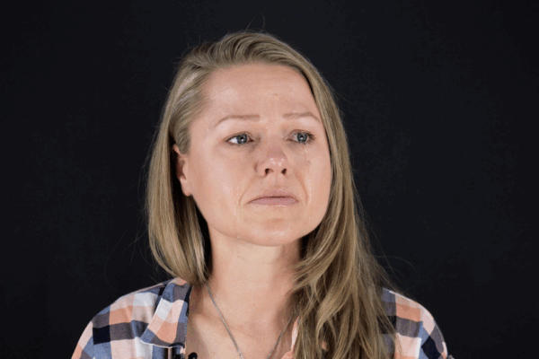 Client, Rebecca Leavitt. She is a white woman with blonde hair, blue eyes, and is wearing a blue, pink, and white checkered collared shirt. She is looking off to the side and has tears down her face.