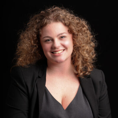 Headshot of Hannah. She is a white woman with light brown curly hair. She is wearing a grey bloue and black blazer.