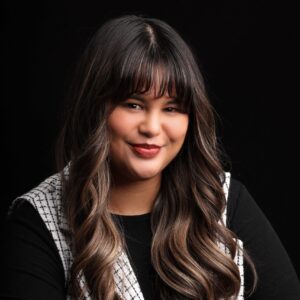 Headshot of Jordyn. She is a brown woman with bangs and highlights. She is wearing a black and white vest and a black long sleeve shirt.