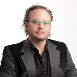 Headshot of Jacob. He is a white male with glasses and is wearing a black and grey blazer with a black collared shirt.