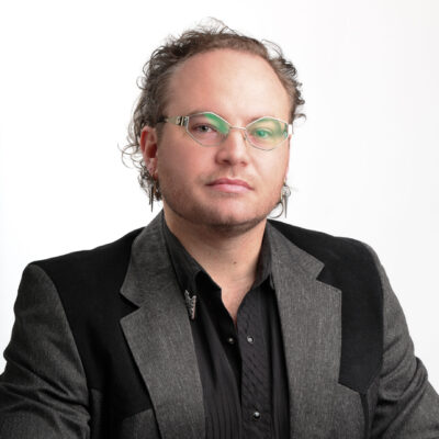 Headshot of Jacob. He is a white male with glasses and is wearing a black and grey blazer with a black collared shirt.