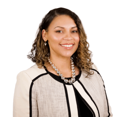 Woman with shoulder-length curly hair, wearing a cream and black jacket with a pearl necklace, smiling in front of a white background.