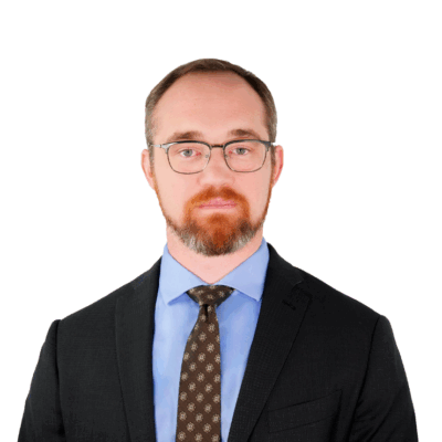 Man with short hair and a beard, wearing glasses, a dark suit, light blue shirt, and patterned tie, facing forward with a neutral expression.
