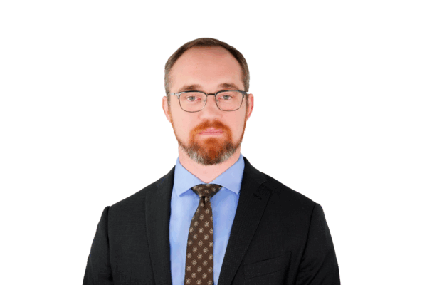 Man with short hair and a beard, wearing glasses, a dark suit, light blue shirt, and patterned tie, facing forward with a neutral expression.