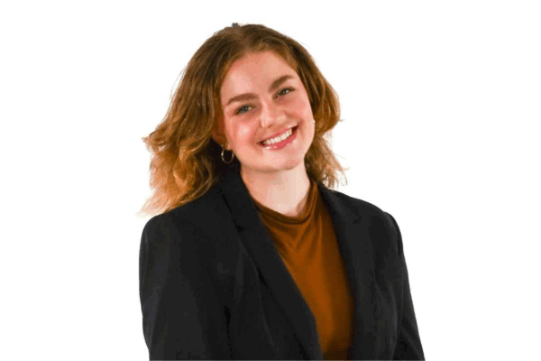 Woman with shoulder-length wavy hair, wearing hoop earrings, a rust-colored top, and a black blazer, smiling in front of a white background.