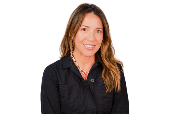 Woman with long wavy hair, wearing a black zip-up jacket and a chunky chain necklace, smiling against a white background.