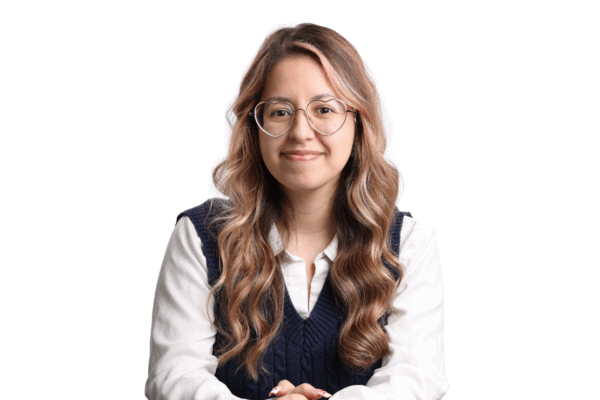 Woman with long wavy hair and light pink highlights, wearing large heart-shaped glasses, a white blouse, and a navy sweater vest, smiling in front of a white background.