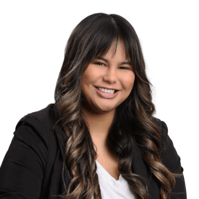 Woman with long, curled dark brown hair and bangs, wearing a black blazer and white blouse, smiling brightly in front of a white background.